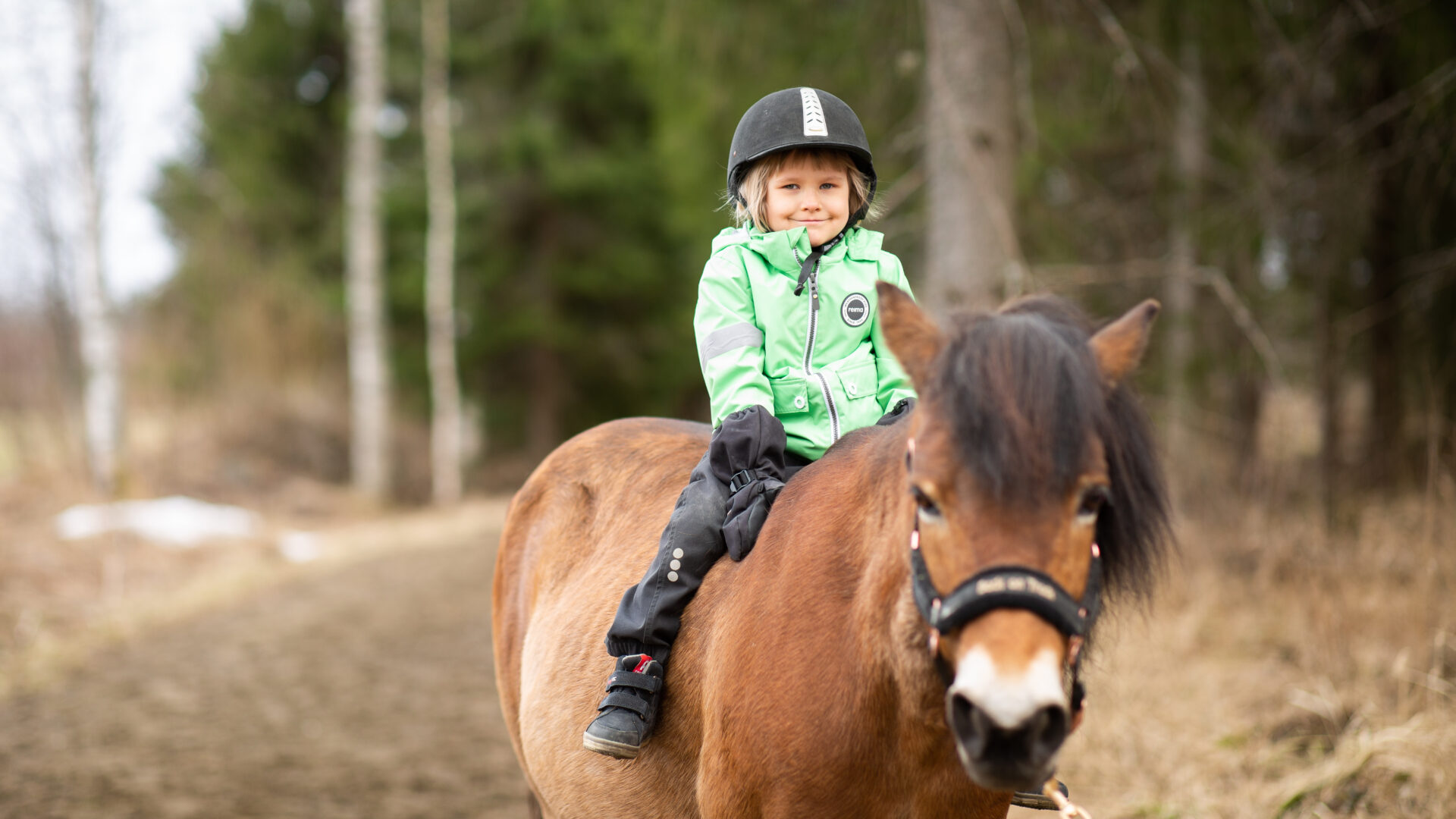 Tyttö istuu hevosen selässä metsäpolulla.