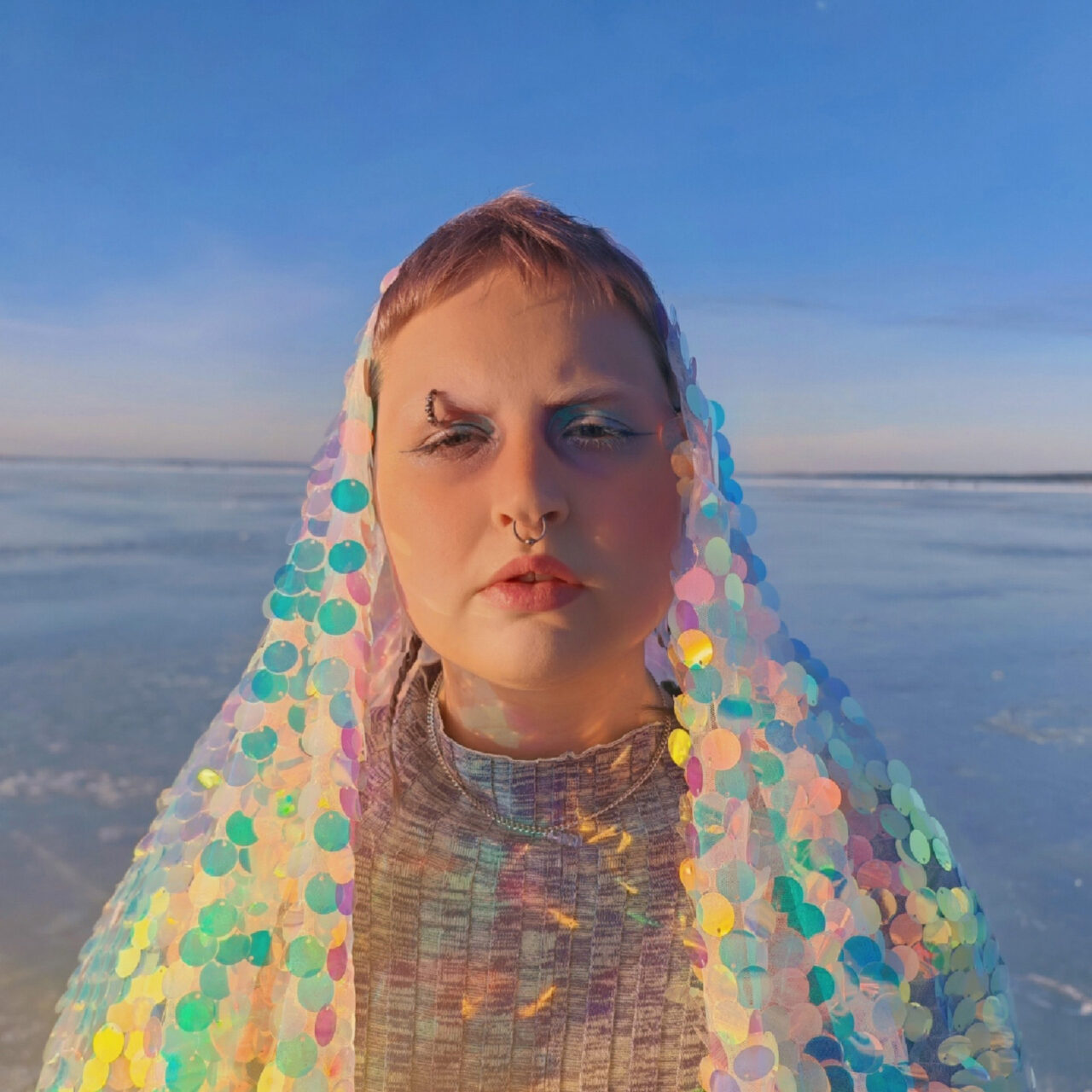 Nuorella naisella on päällään huntumainen vaate, jossa on värikkäitä paljetteja. Taustalla on jäätynyt järvi.