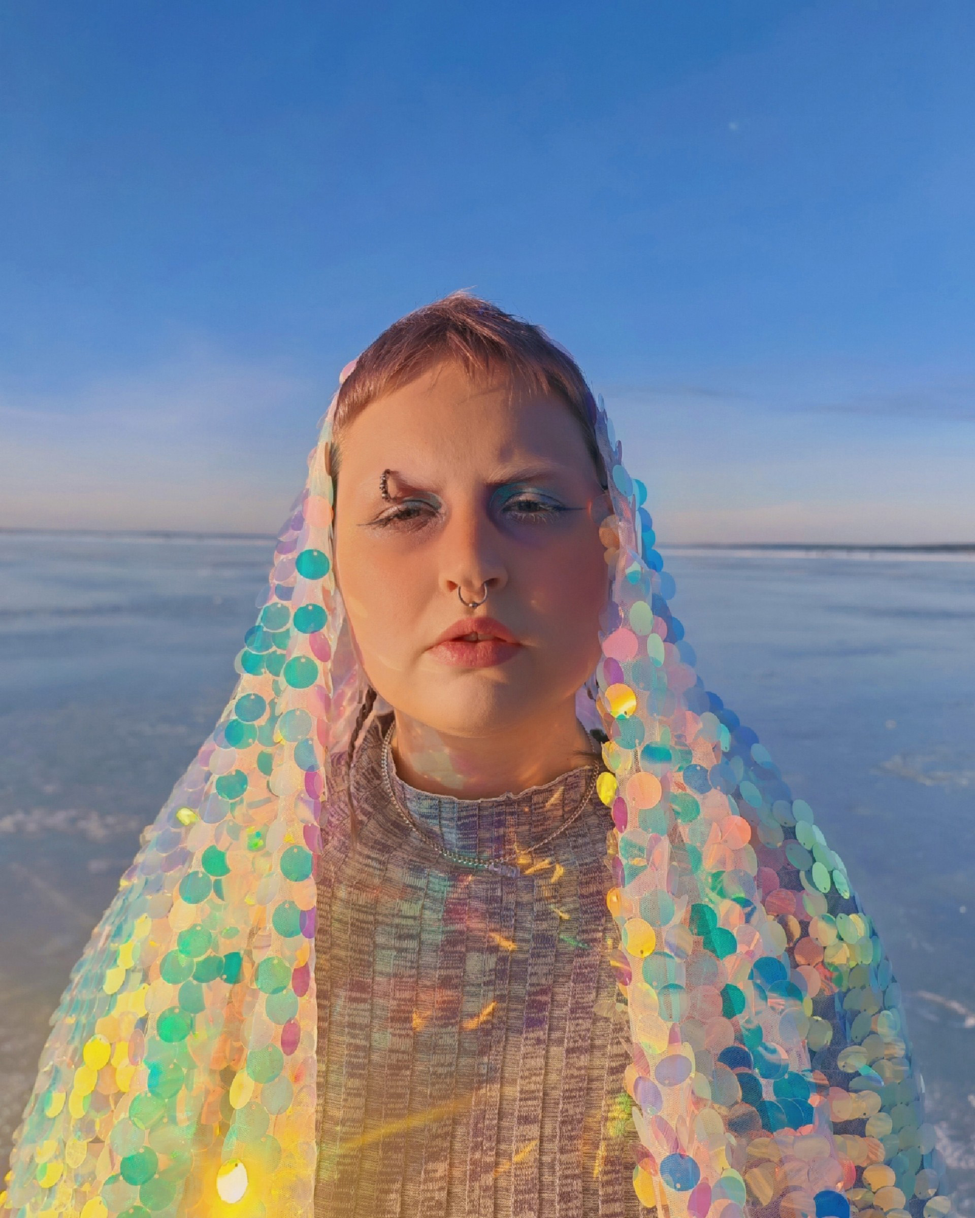 Nuorella naisella on päällään huntumainen vaate, jossa on värikkäitä paljetteja. Taustalla on jäätynyt järvi.