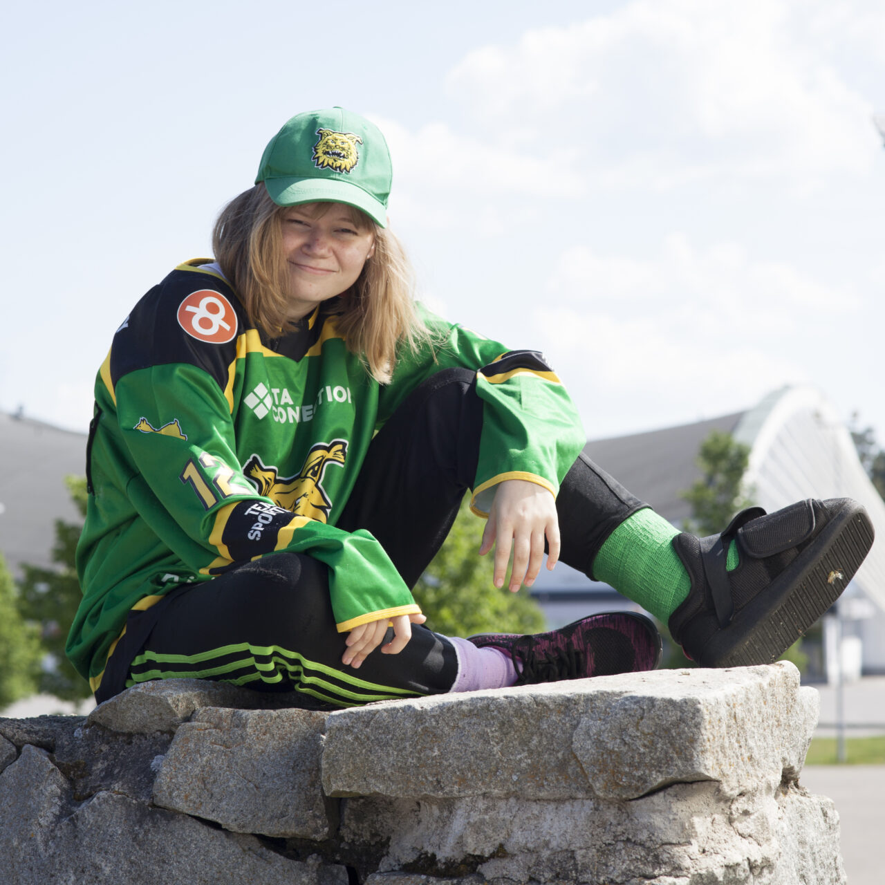 Tyttö istuu kiviaidan päällä. Hänellä on päällään vihreä Ilves-joukkueen pelipaita ja lippis.