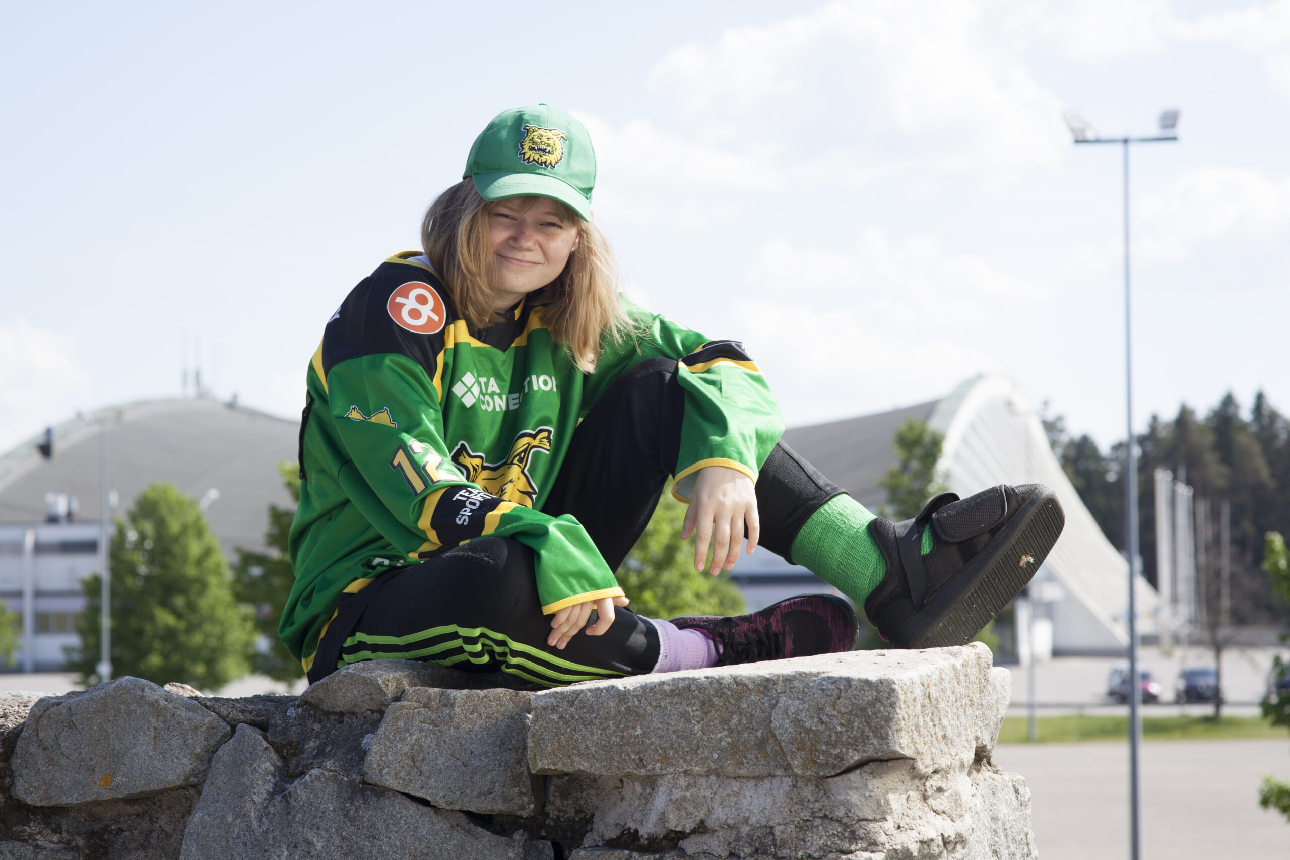 Tyttö istuu kiviaidan päällä. Hänellä on päällään vihreä Ilves-joukkueen pelipaita ja lippis.