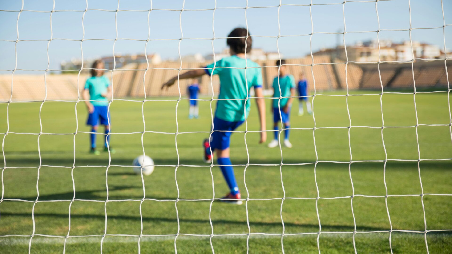 Lapset pelaavat jalkapallokentällä, joka on kuvattu maaliverkon takaa.