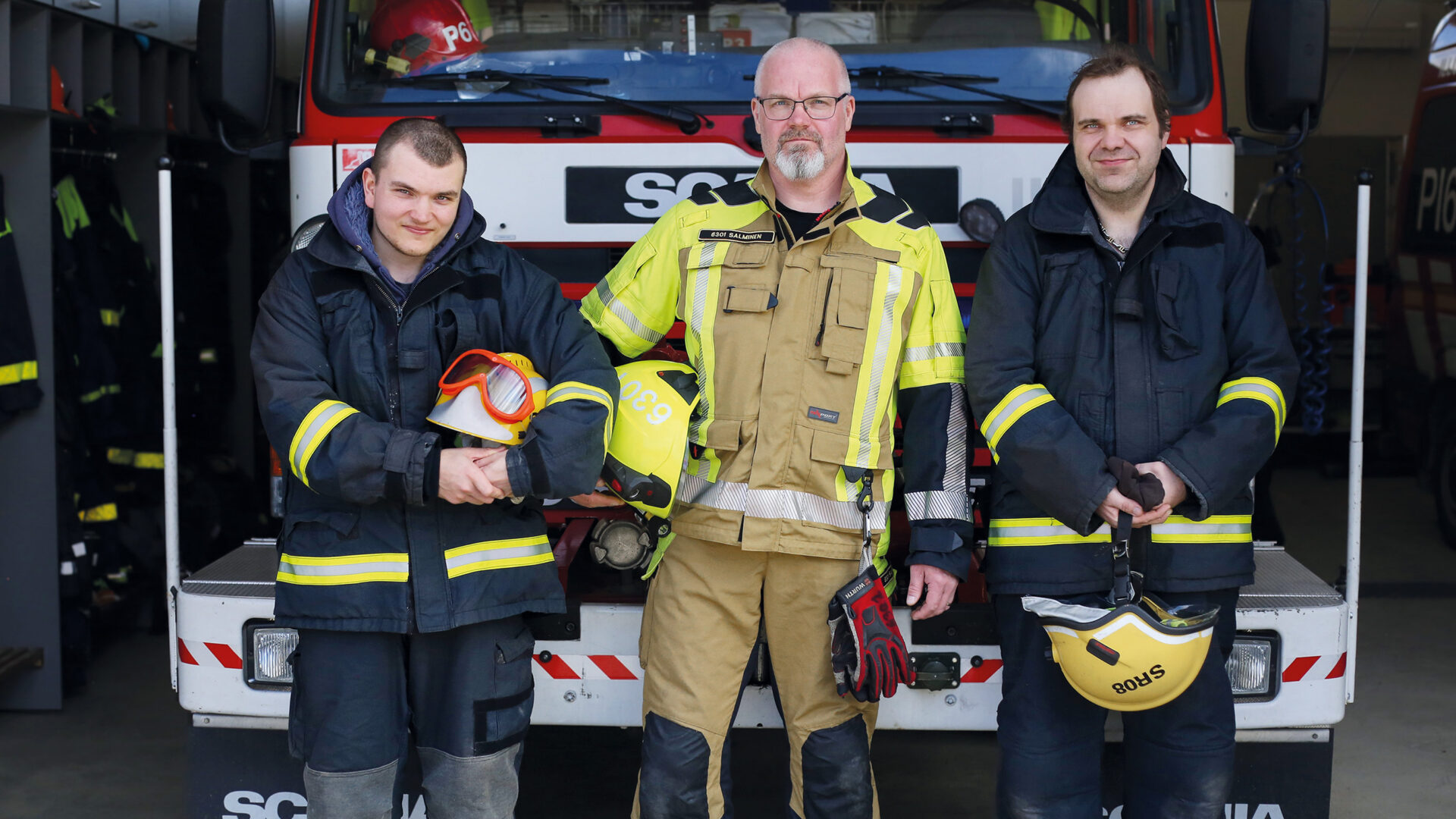 Kolme miestä palomiehen varusteissa paloauton edessä.