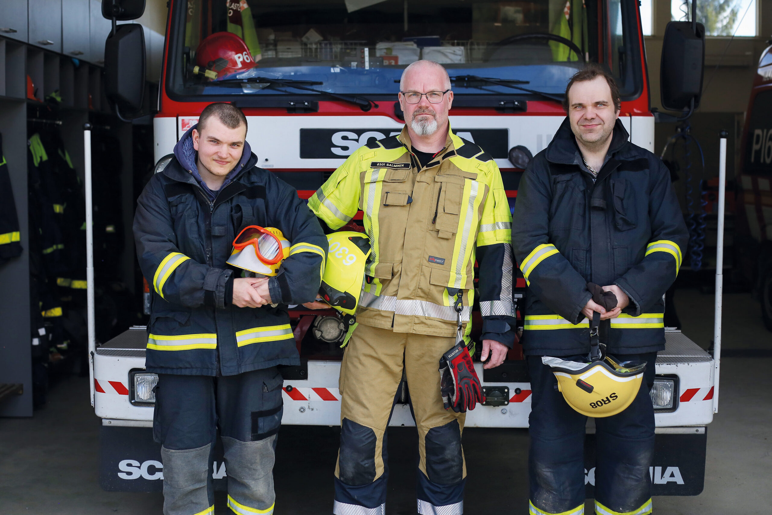 Kolme miestä palomiehen varusteissa paloauton edessä.