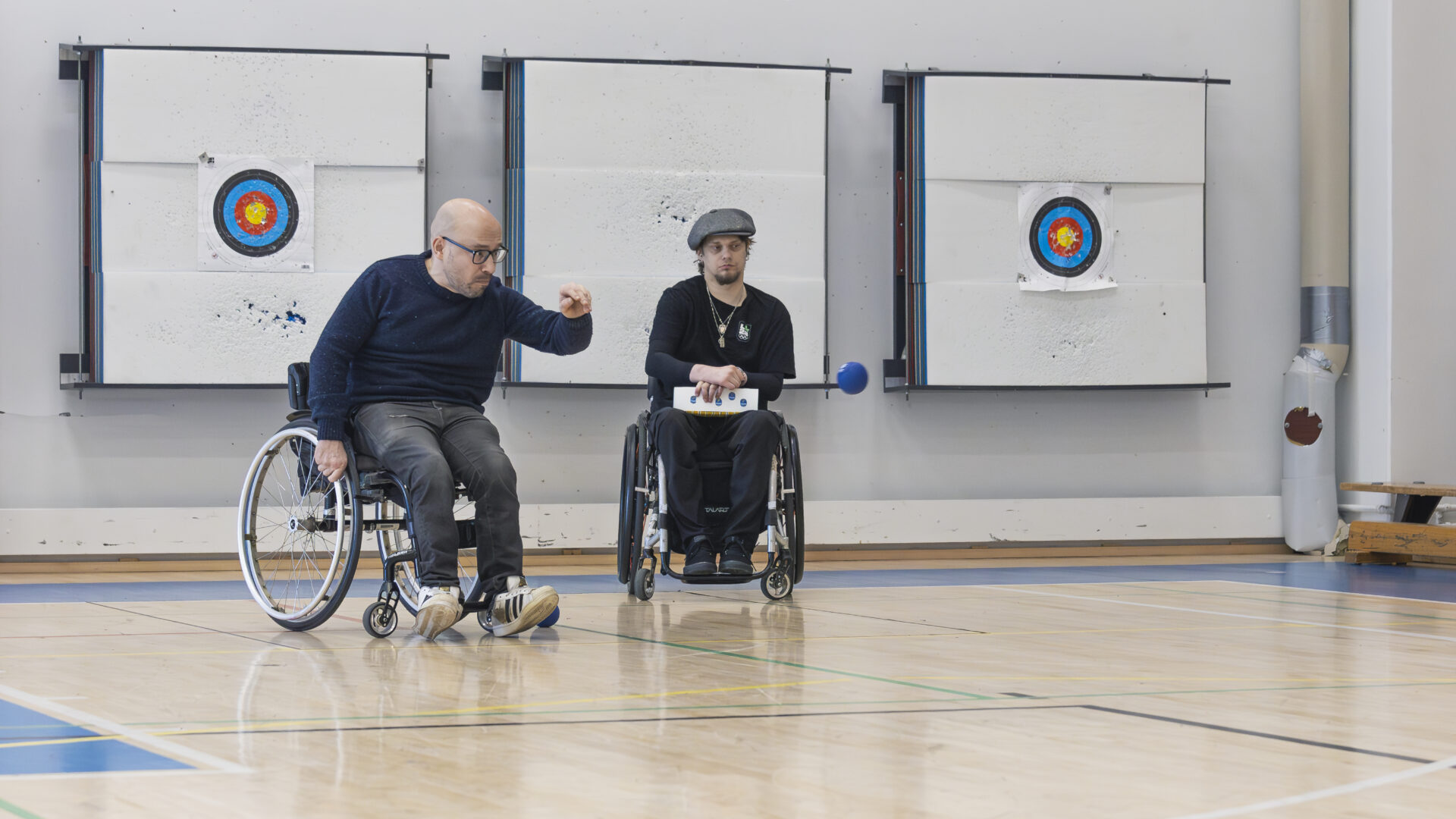 Kaksi miestä pelaa bocciaa pyörätuolista. Mies heittää sinistä palloa.