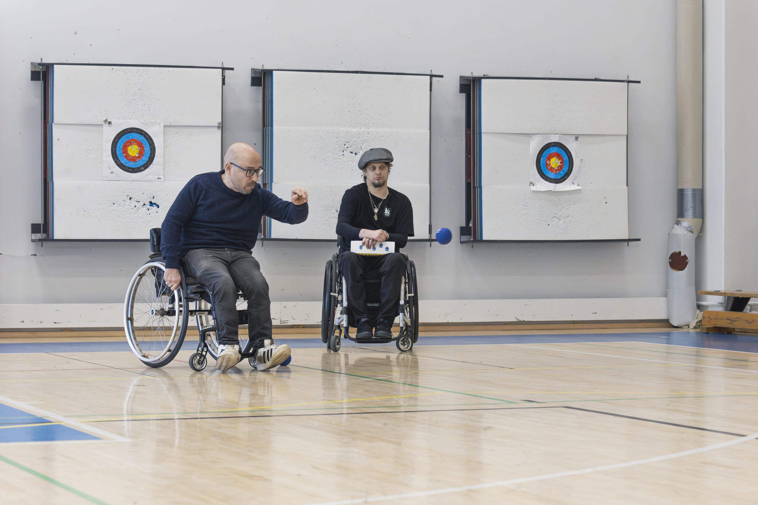 Kaksi miestä pelaa bocciaa pyörätuolista. Mies heittää sinistä palloa.