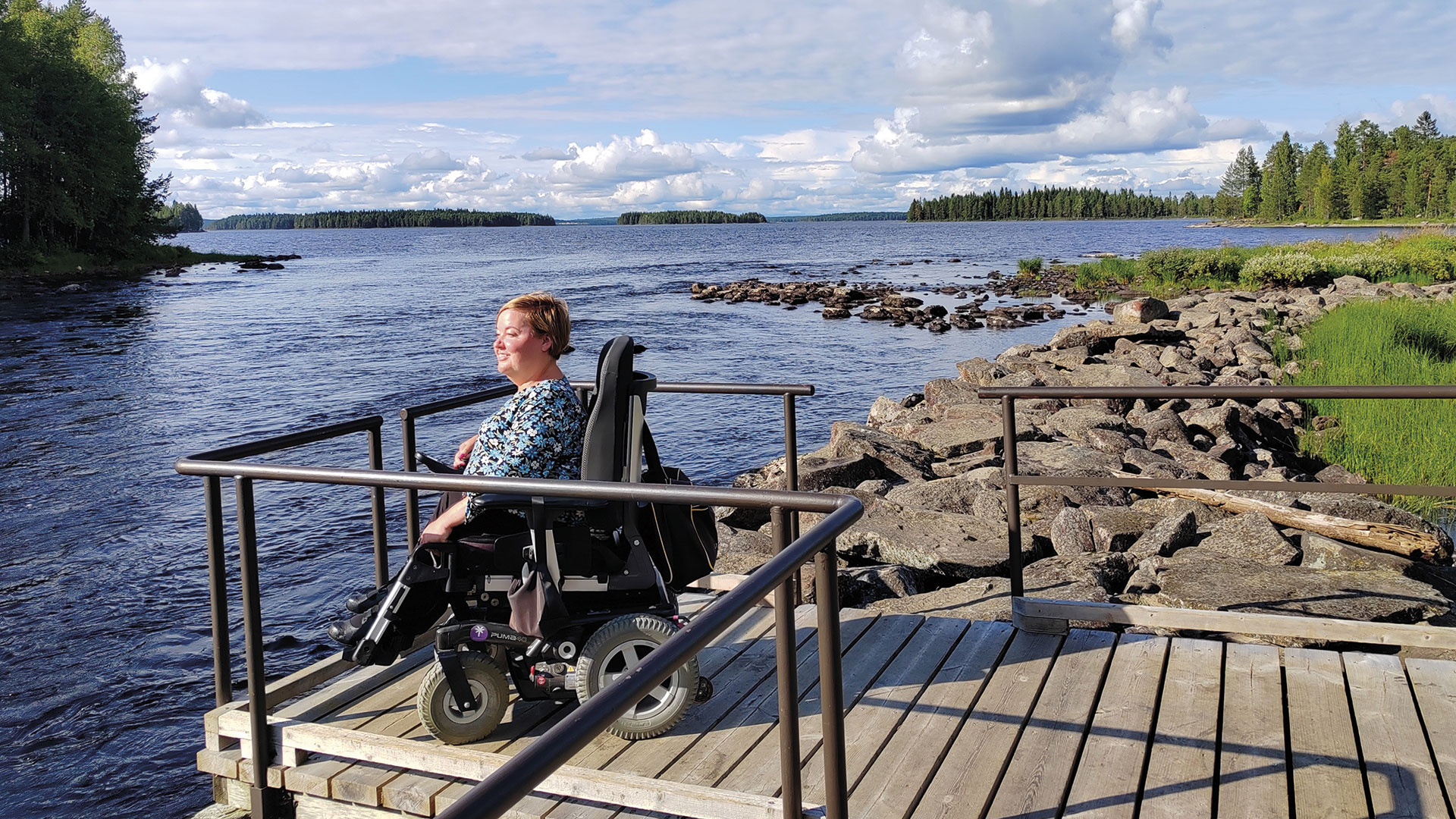 Sanna Kalmari istuu pyörätuolissa laiturilla ja katsoo järvimaisemaa.