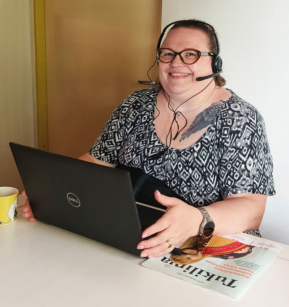 Elina Miettinen pöydän ääressä kuulokkeet korvilla ja kannettava tietokone edessään.