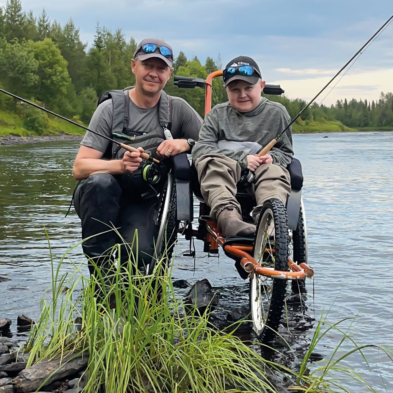 Jere Huovinen istuu kalastukseen sopivassa pyörätuolissa rantavedessä virveli kädessä. Vierellä isä Teemu Huovinen.