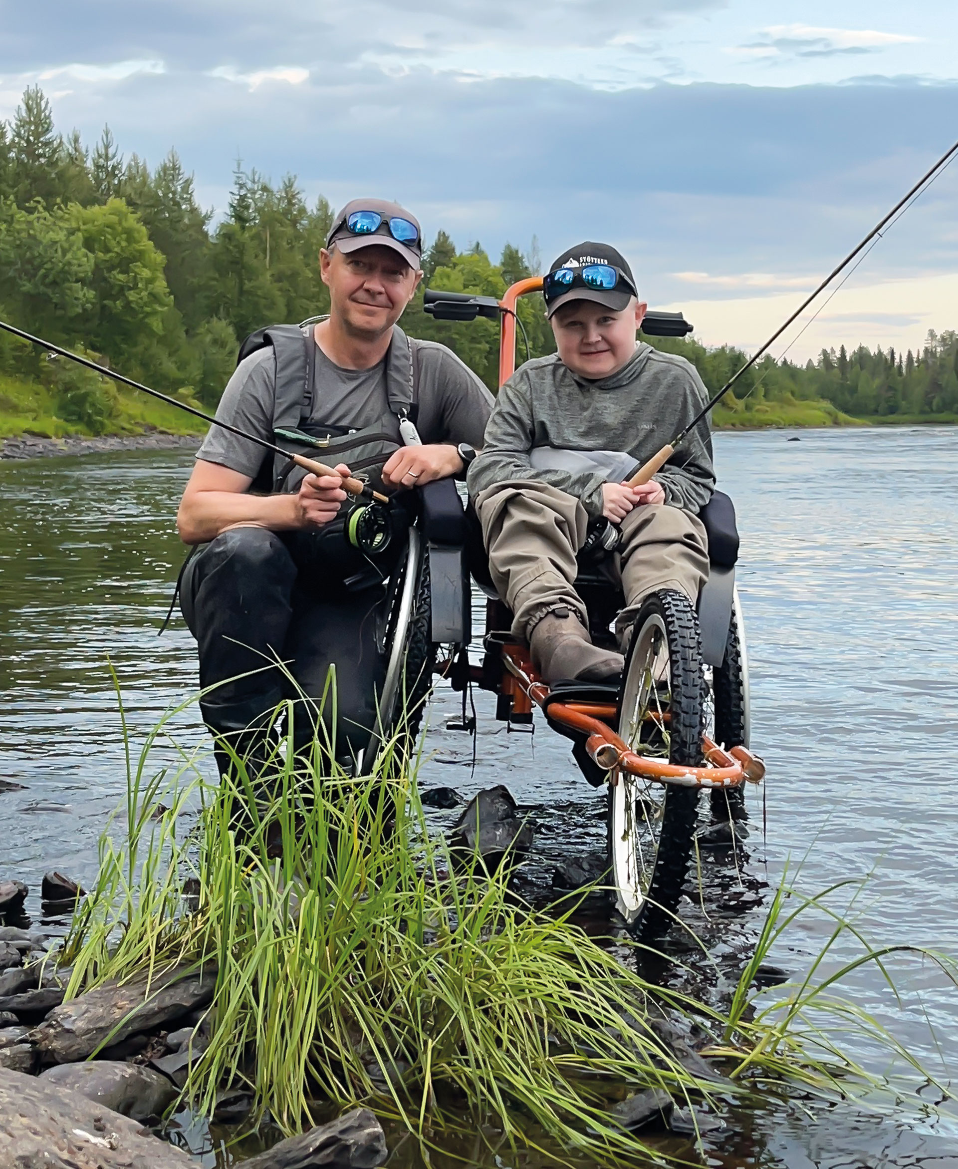 Jere Huovinen istuu kalastukseen sopivassa pyörätuolissa rantavedessä virveli kädessä. Vierellä isä Teemu Huovinen.